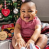 baby, book, child, christmas_tree, cute, decorations, festive, happy, holiday, indoor, infant, lights, ornaments, person, plaid_blanket, red_clothing, sitting, smiling, sock, toy