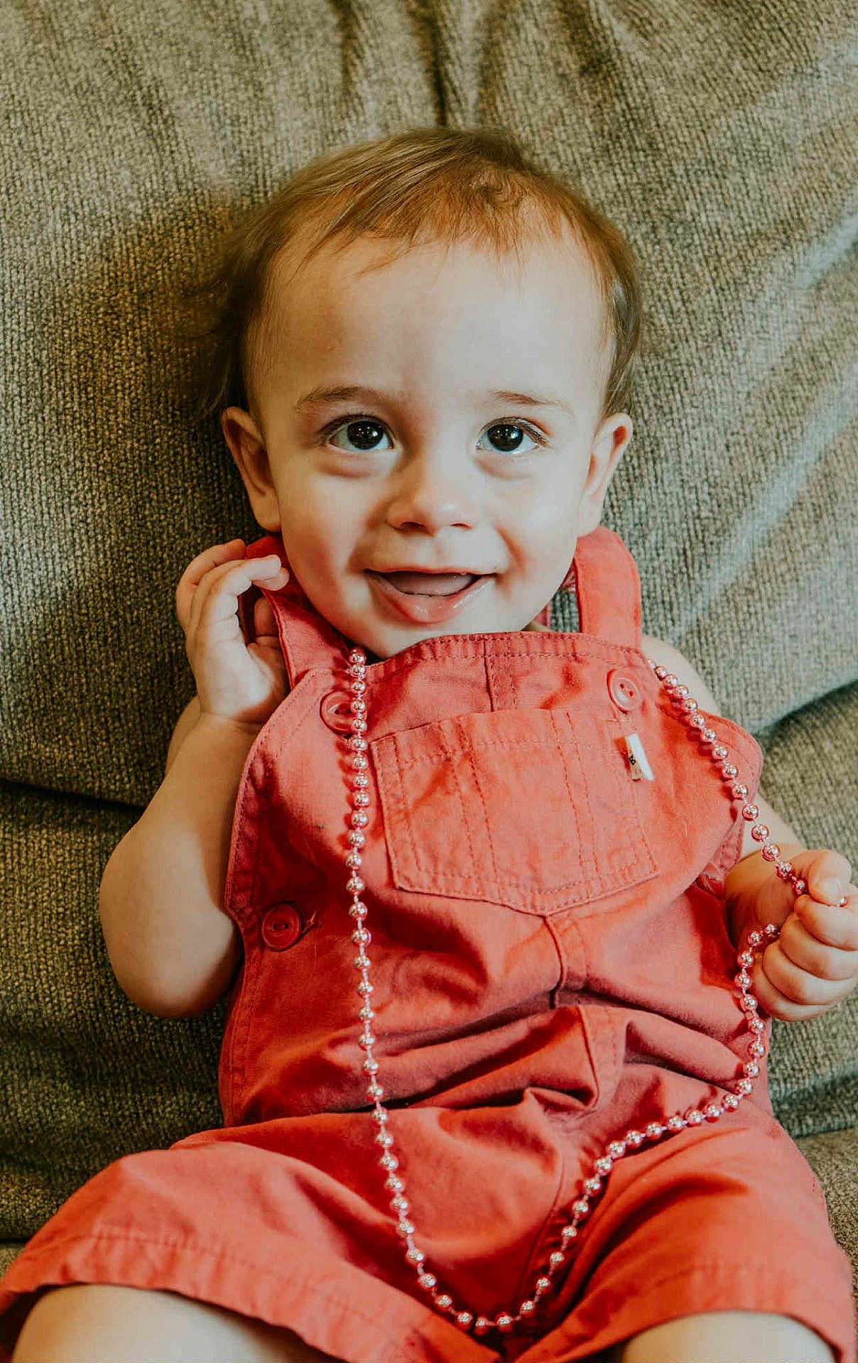 Beckham is registered to the contest to win money with this photo: toddler, child, smiling, overalls, orange_clothing, bead_necklace, sitting, couch, happy, face, hands, indoor, portrait, cute, young_child, clothing, person, baby, expression, furniture