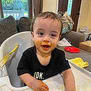 Beckham is registered to the contest to win money with this photo: toddler, child, high_chair, messy, food, sauce, face, hands, black_shirt, indoor, living_room, window, curtains, sofa, plate, table, bag, yellow_cloth, door, happy