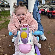 Nova joined the competition — help win amazing prizes! toddler, child, pink_jacket, unicorn_toy, outdoor, toy, playground, adult_hands, jeans, sneakers, grass, dirt, toy_cars, fence, blue_sky, person, cute, face, handlebars, play