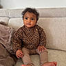 child, toddler, curly_hair, leopard_print, brown_clothing, beige_couch, blanket, indoor, casual, sitting, barefoot, portrait, cute, young_child, cozy, home, furniture, relaxed, small_hands, curious_eyes