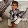 baby, child, curly_hair, sitting, couch, blanket, pillow, indoors, clothing, checkered, pants, barefoot, cute, smiling, hand, face, person, cozy, soft_fabric, relaxed