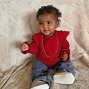 Malika a rejoint le concours — aidez-le/la à gagner de superbes lots ! toddler, child, red_clothing, jeans, chain, curly_hair, sitting, blanket, indoor, footwear, cute, smiling, portrait, baby, fashion, happy, comfortable, casual, young_child, playful