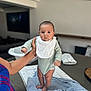 Devon joined the competition — help win amazing prizes! baby, infant, onesie, bib, hand, towel, table, indoor, person, support, standing, soft, blurred_background, living_room, furniture, adult, skin, face, eyes, clothing