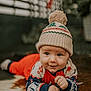 baby, infant, hat, pom_pom, red_clothing, patterned_sleeves, fur_rug, lying_down, blue_eyes, smiling, cute, indoors, portrait, child, winter_clothing, cozy, warm, close_up, face, happy