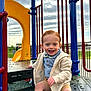 toddler, child, playground, slide, smiling, happy, blue_eyes, bow_tie, jacket, shorts, sneakers, outdoor, play_equipment, daylight, cloudy_sky, fence, metal, plastic, fun, casual_clothing