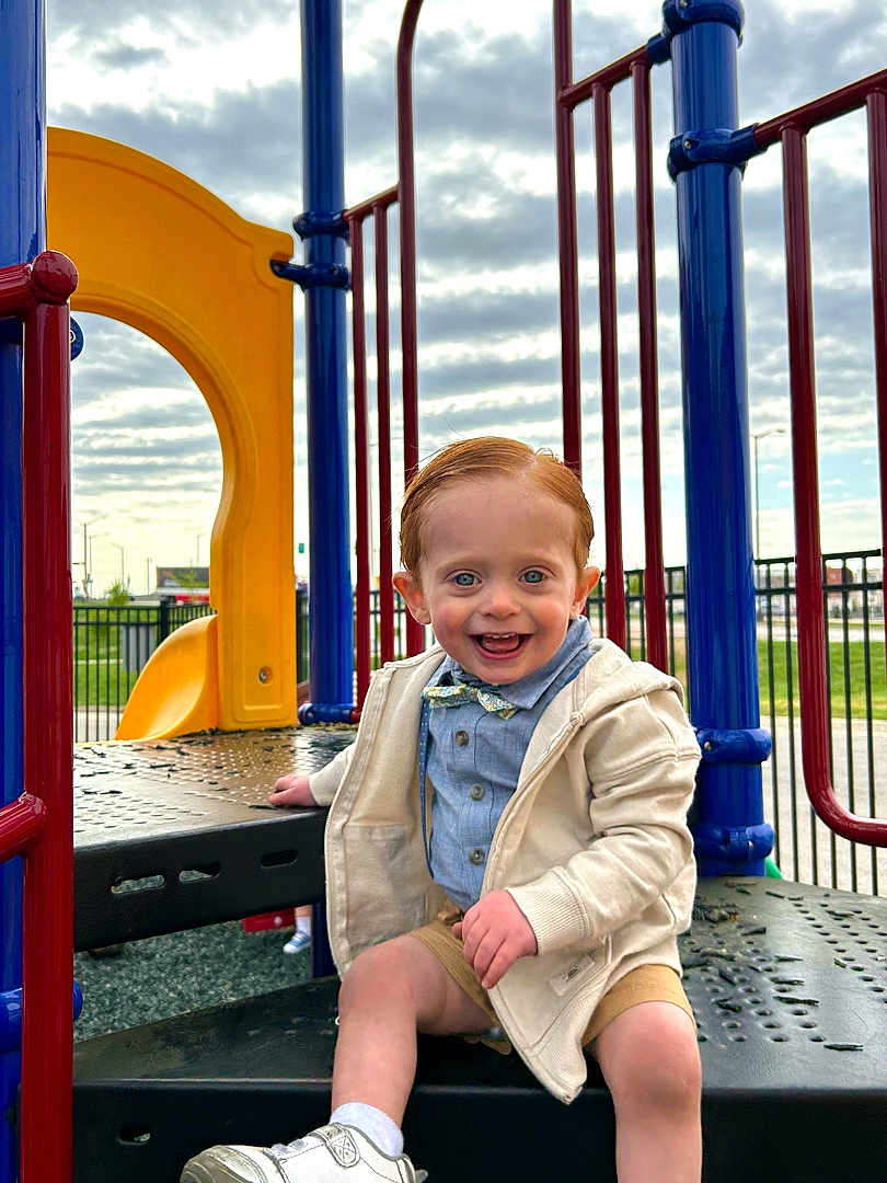 Charlie joined the competition — help win amazing prizes! toddler, child, playground, slide, smiling, happy, blue_eyes, bow_tie, jacket, shorts, sneakers, outdoor, play_equipment, daylight, cloudy_sky, fence, metal, plastic, fun, casual_clothing