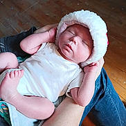Legend is registered to the contest to win money with this photo: baby, newborn, sleeping, hat, white_clothing, arms, person, floor, wooden_floor, gift_wrap, hands, feet, indoors, cozy, peaceful, infant, cute, resting, holding, soft