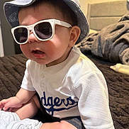 Alexander joined the competition — help win amazing prizes! baby, sunglasses, hat, child, bed, clothing, white_shoes, person, indoors, cute, fashion, denim, portrait, sitting, footwear, headwear, casual, infant, smile, cozy