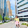 child, girl, urban, cityscape, buildings, skyscraper, sidewalk, pedestal, outdoor, daylight, blue_sky, trees, cars, casual_clothing, playful, smiling, street, modern_architecture, person, young