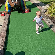 Elianna joined the competition — help win amazing prizes! toddler, child, mini_golf, statue, bear, green_carpet, outdoor, sunny, shadow, building, grass, concrete, pathway, playground, casual_clothing, white_shoes, colorful, fun, daytime, young_child