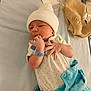 baby, baby_clothes, baby_face, baby_wrists_band, beige_clothing, blue_clothing, cozy, cute, hand_near_mouth, hospital, hospital_bed, infant, lying_down, medical_bottle, newborn, onesie, sleepy, small_hands, soft_fabric, white_hat