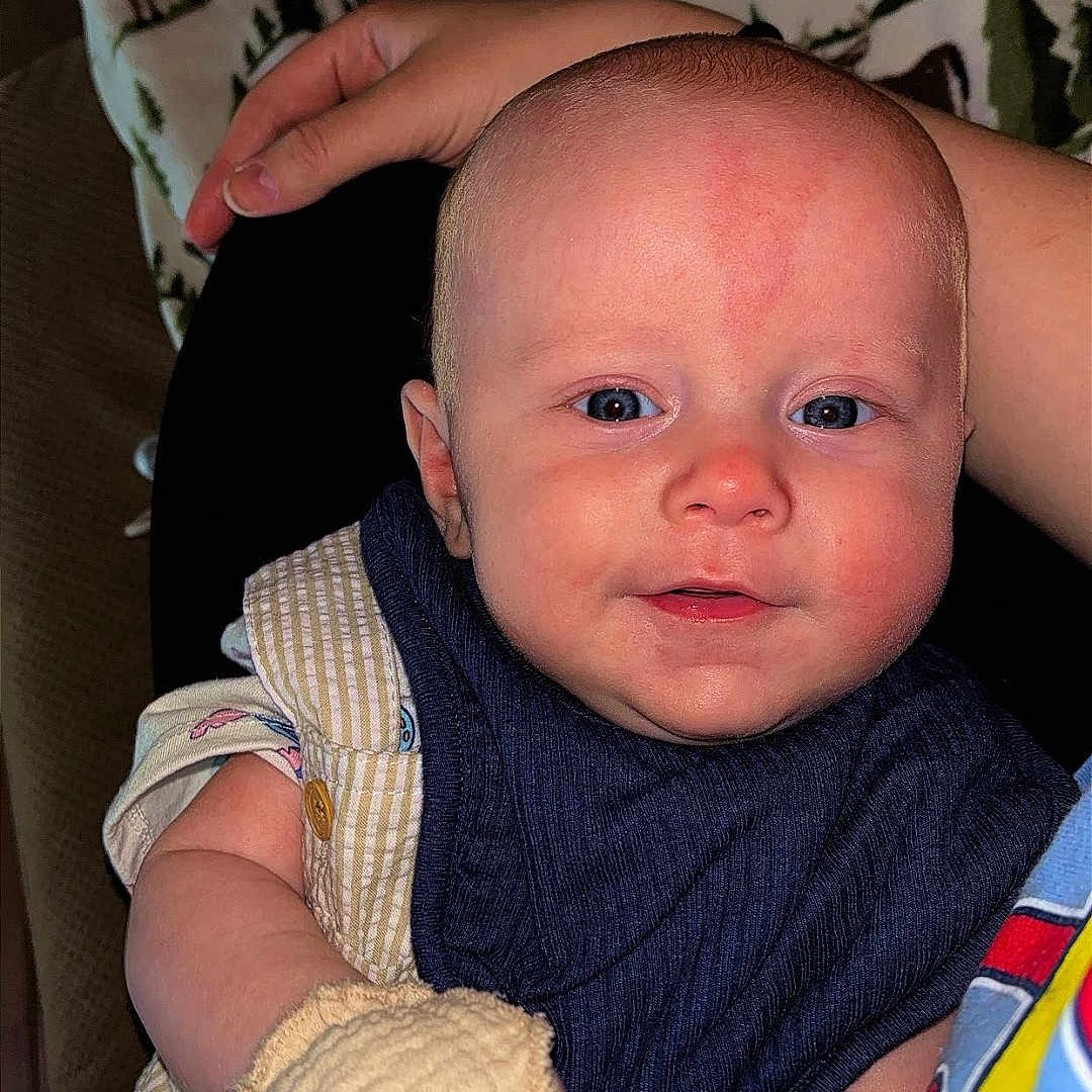 Asher is registered to the contest to win money with this photo: baby, infant, mittens, blue_eyes, bib, blanket, hand, person, clothing, face, smiling, indoor, portrait, cute, skin, head, sitting, comfort, young_child, pattern