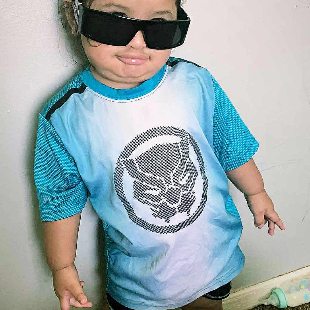 Nooreeann joined the competition — help win amazing prizes! child, sunglasses, blue_shirt, black_shorts, barefoot, carpet, baby_bottle, standing, wall, hair_bun, smile, indoor, person, toddler, funny_pose, casual_clothing, light_skin, shorts, toy, cute