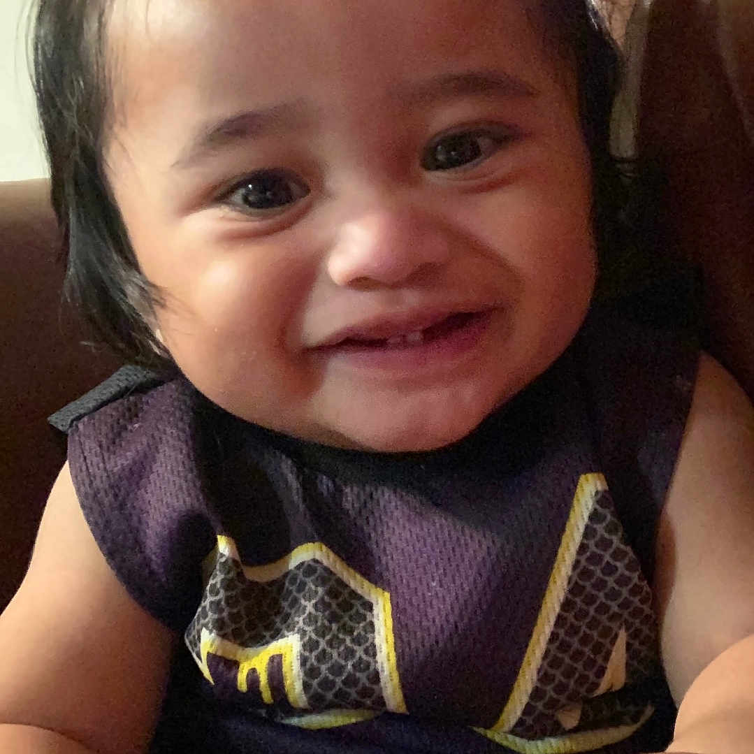 Gm joined the competition — help win amazing prizes! toddler, child, smile, face, hair, pony_tail, sleeveless_shirt, patterned_clothing, brown_background, skin, hand, happy, indoors, portrait, cute, person, young_child, seated, closeup, expression