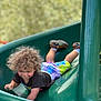 active, casual_clothing, child, colorful_clothing, curly_hair, daylight, fun, green_slide, happy, nature_background, outdoor, play, playground, shoes, slide, smile, socks, summer, toddler, young_child