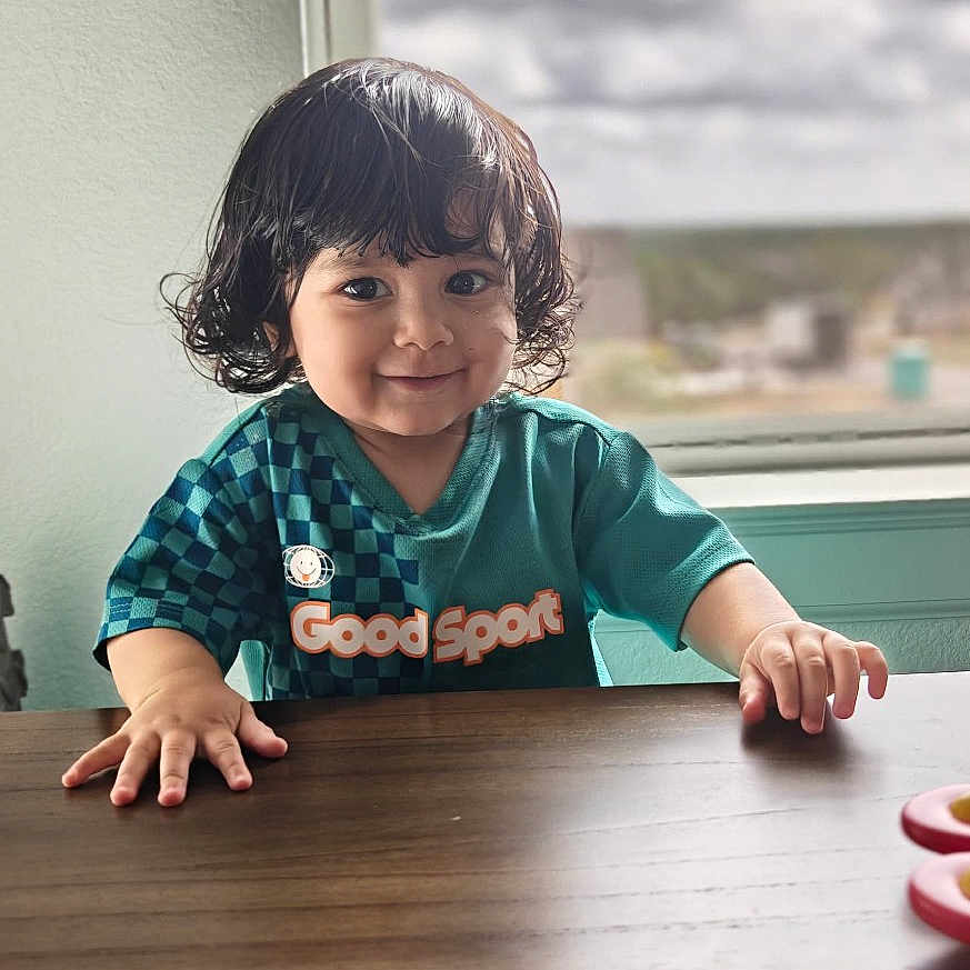Saumya Sethna is registered to the contest to win money with this photo: child, toddler, smiling, curly_hair, teal_shirt, good_sport, table, wooden_table, indoor, window, cloudy_sky, hands, face, portrait, happy, person, young_child, curious, seated, natural_light
