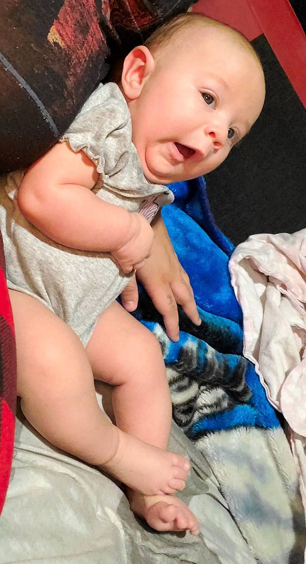 Arizona Rose is registered to the contest to win money with this photo: baby, bedding, blanket, blue_blanket, child, clothing, cozy, eyes, face, feet, gray_outfit, hand, happy, indoors, infant, lying_down, mouth, portrait, red_background, smiling