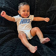 Azai is registered to the contest to win money with this photo: baby, child, smiling, happy, nike, onesie, infant, sitting, black_blanket, cozy, cute, portrait, baby_boy, barefoot, indoor, young_child, playful, expression, clothing, relaxed