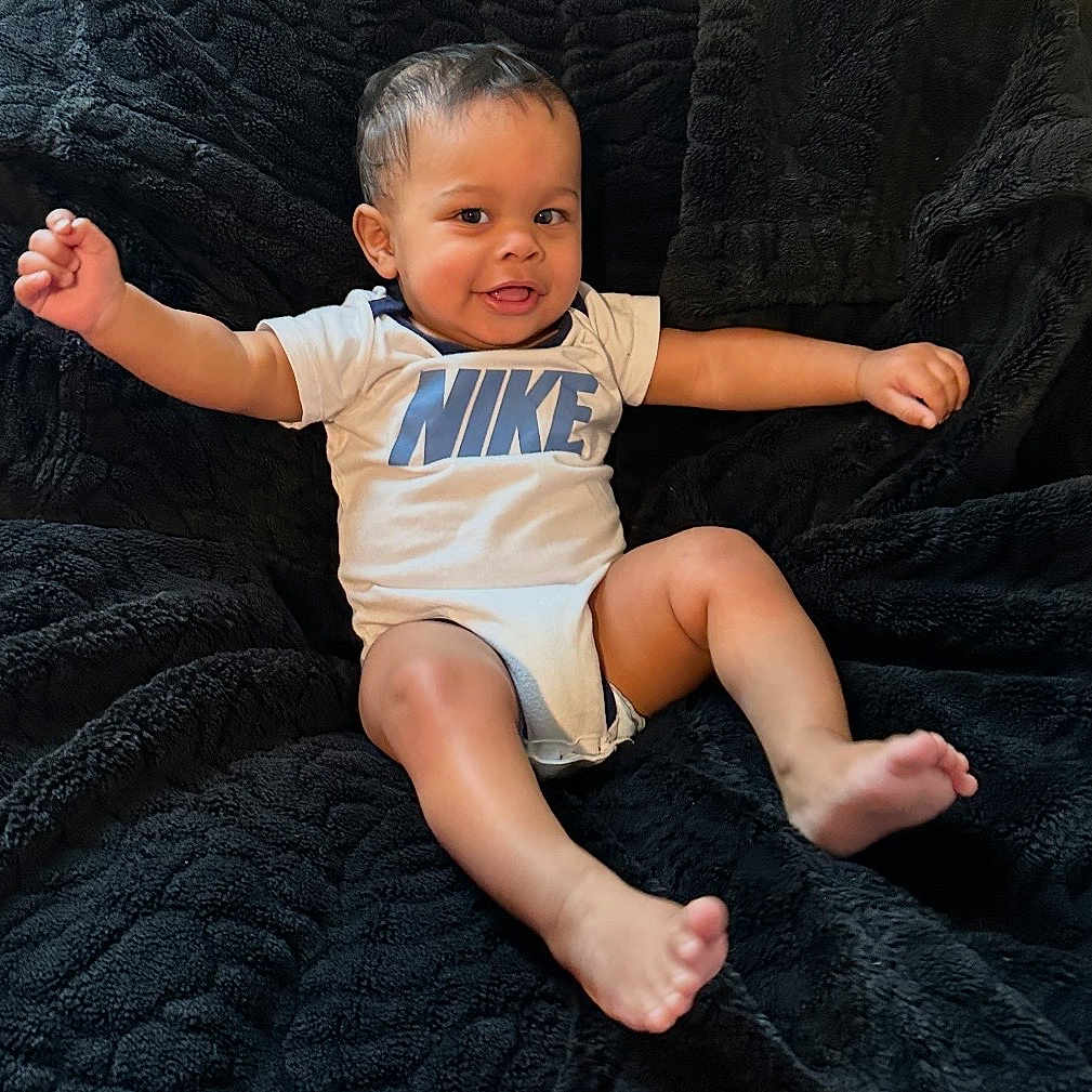 Azai is registered to the contest to win money with this photo: baby, child, smiling, happy, nike, onesie, infant, sitting, black_blanket, cozy, cute, portrait, baby_boy, barefoot, indoor, young_child, playful, expression, clothing, relaxed