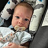 baby, infant, child, portrait, face, eyes, pacifier, blanket, car_seat, safety_harness, seated, indoor, cozy, awake, looking_at_camera, brown_hair, blue_clothing, patterned_fabric, hand, close_up