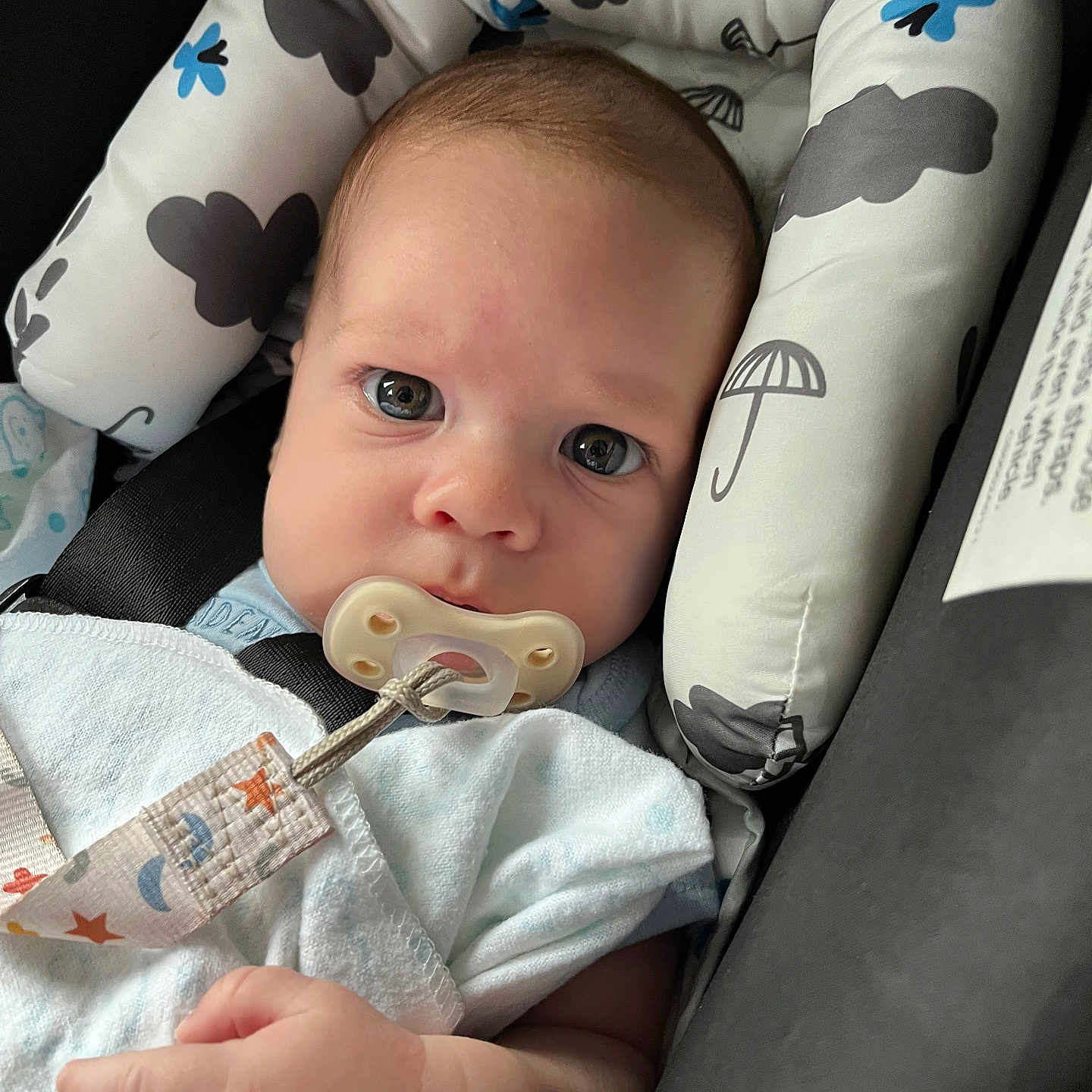 Wyatt is registered to the contest to win money with this photo: baby, infant, child, portrait, face, eyes, pacifier, blanket, car_seat, safety_harness, seated, indoor, cozy, awake, looking_at_camera, brown_hair, blue_clothing, patterned_fabric, hand, close_up
