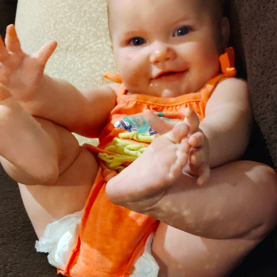 Maci is registered to the contest to win money with this photo: baby, child, couch, cozy, cute, diaper, face, foot, furniture, hand, happy, indoors, infant, orange_clothing, person, plush, relaxed, sitting, skin, smiling