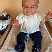 Mathéïs participe au concours pour gagner de l'argent avec cette photo : baby, blonde_hair, blue_ball, changing_pad, child, cute, dark_pants, expression, face, hands, indoor, light_skin, playful, sitting, smiling, socks, toddler, toy, white_tshirt, wooden_furniture