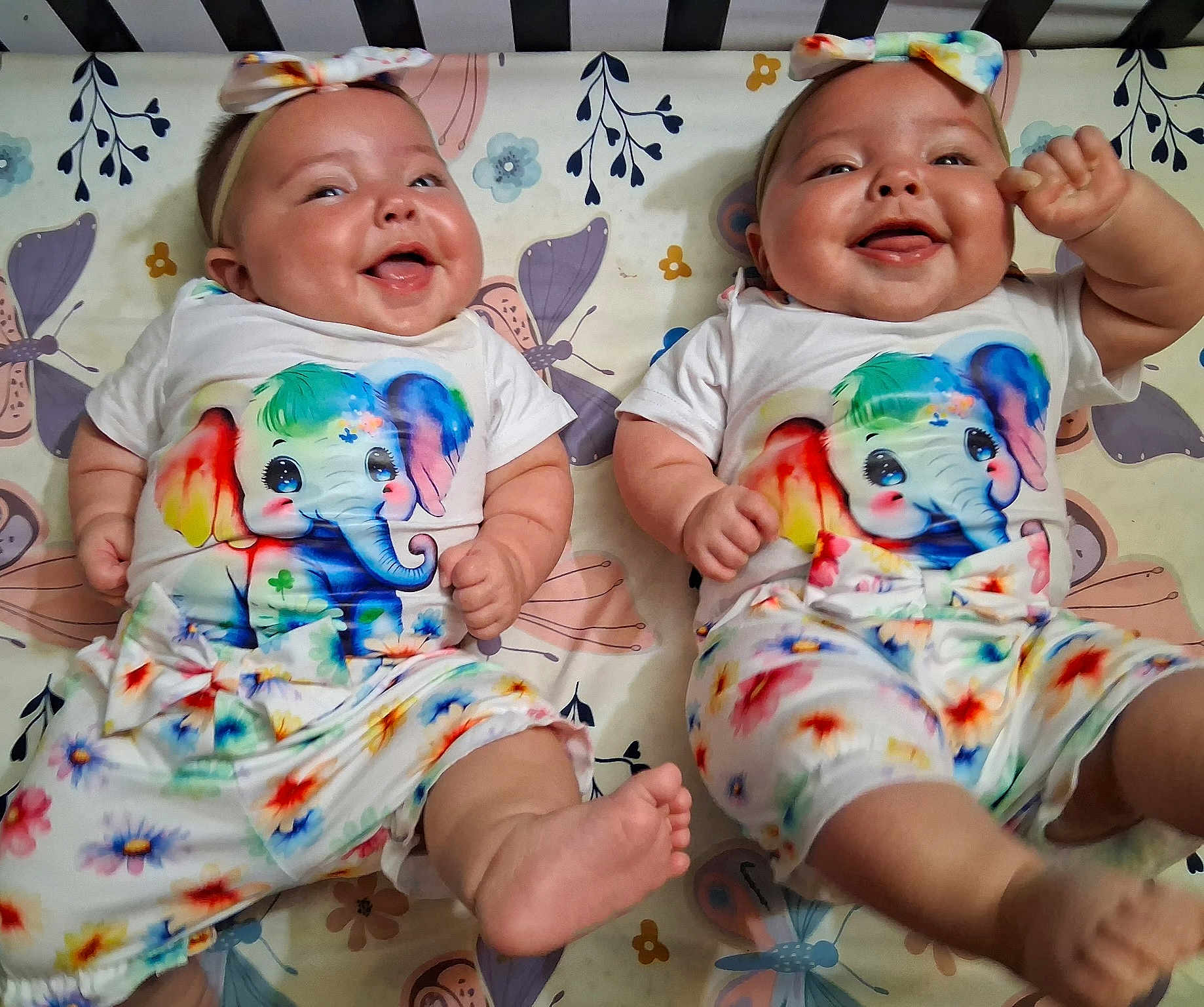 Juanita And Regina joined the competition — help win amazing prizes! baby, twins, infant, smiling, lying_down, headband, colorful_clothing, elephant_print, floral_pattern, cute, happy, chubby, indoors, crib, person, toddler, playful, joyful, apparel, portrait