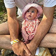 Lennie is registered to the contest to win money with this photo: baby, child, person, hat, pink, checkered, cherry_pattern, outdoor, grass, adult, hands, legs, sitting, casual_clothing, sneakers, wristband, nature, daylight, curious_expression, closeup