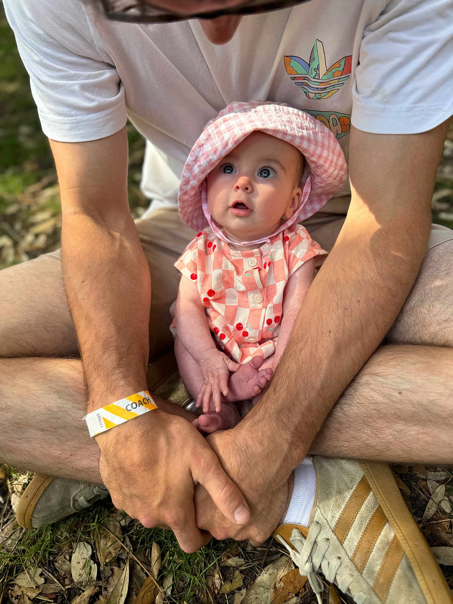 Lennie is registered to the contest to win money with this photo: baby, child, person, hat, pink, checkered, cherry_pattern, outdoor, grass, adult, hands, legs, sitting, casual_clothing, sneakers, wristband, nature, daylight, curious_expression, closeup