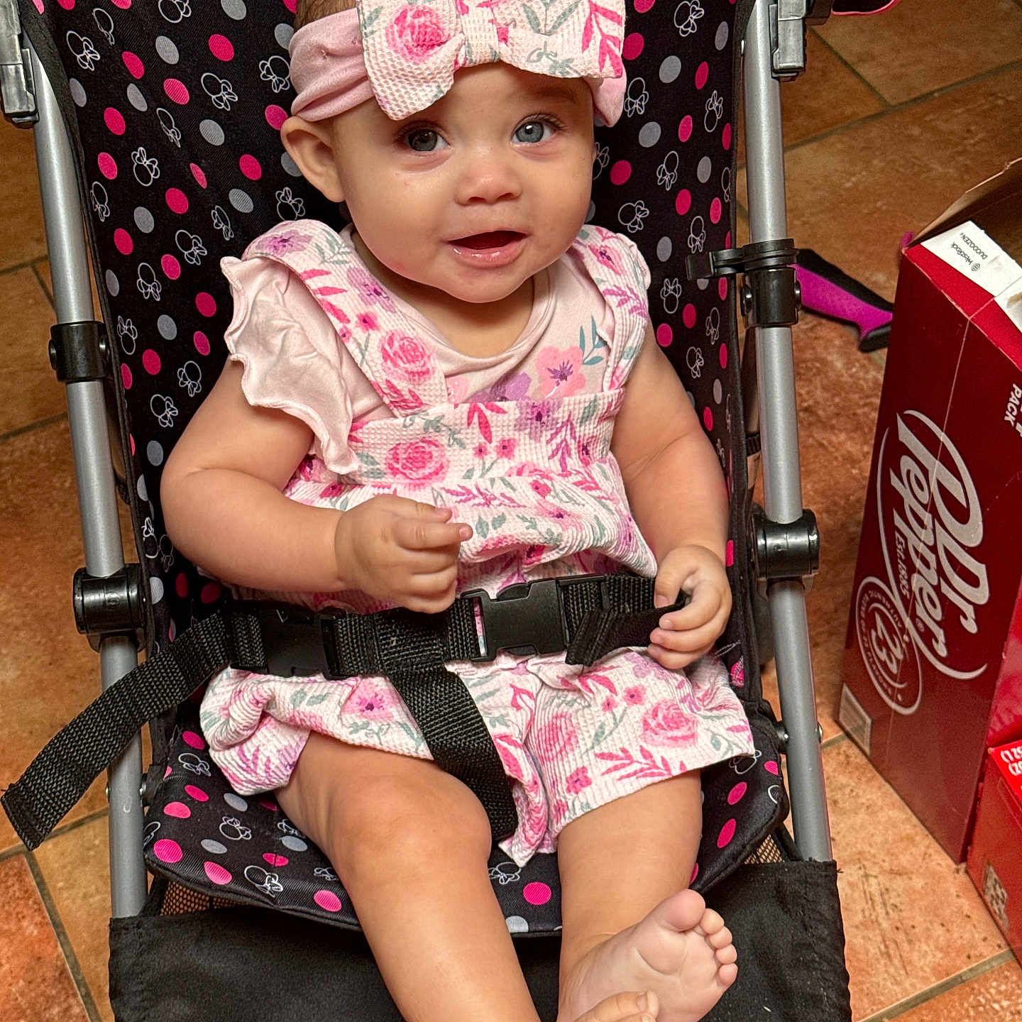 Anastasia is registered to the contest to win money with this photo: baby, box, child, cute, expression, floor_tiles, floral_dress, footwear, happy, headband, indoor, pattern, person, pink, polka_dot, portrait, seatbelt, smiling, stroller, toddler