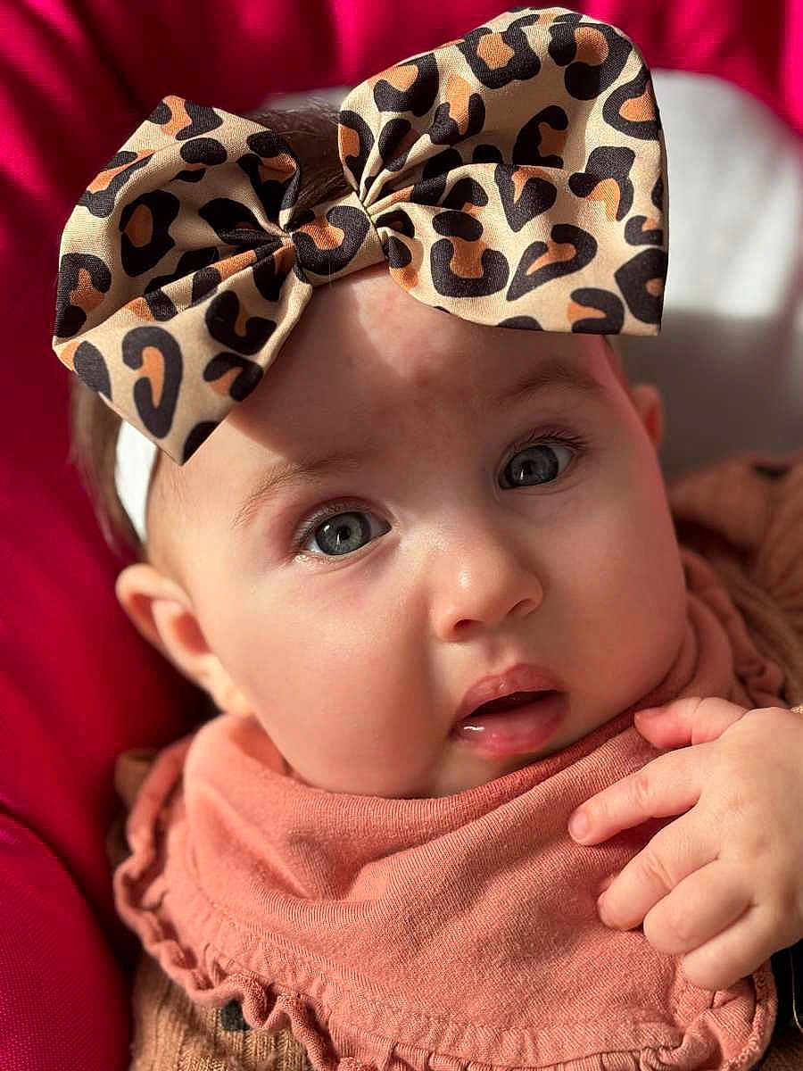 Anaë a rejoint le concours — aidez-le/la à gagner de superbes lots ! baby, infant, headband, bow, face, eyes, cheeks, hand, mouth, portrait, close_up, pink_background, clothing, bib, soft_skin, adorable, eyelashes, headwear, fabric, seat