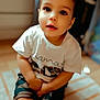 toddler, child, sitting, white_tshirt, camo_shorts, indoor, rug, curious, face, portrait, young_child, short_hair, hands_together, floor, natural_light, home, casual_clothing, smiling, cute, closeup