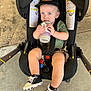 toddler, child, baby, stroller, sippy_cup, hat, cap, black_shoes, sneakers, concrete_floor, child_seat, sitting, straps, clothing, person, footwear, outdoor, expression, hydration, casual