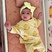 Ayla is registered to the contest to win money with this photo: baby, infant, yellow_clothing, headband, bow, mattress, crib, cute, child, indoors, patterned_clothing, lying_down, pink, decor, expression, small_child, face, person, raised_arm, soft_lighting