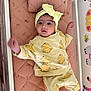 baby, infant, yellow_clothing, headband, bow, mattress, crib, cute, child, indoors, patterned_clothing, lying_down, pink, decor, expression, small_child, face, person, raised_arm, soft_lighting
