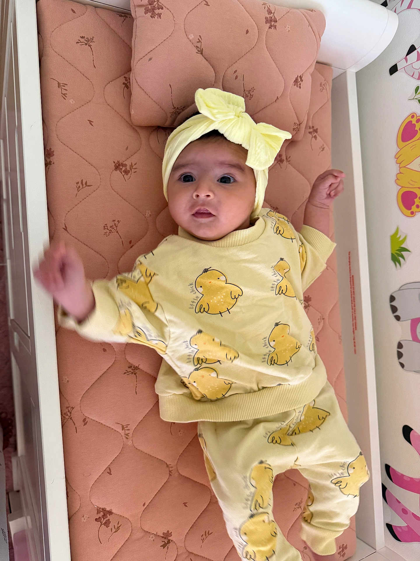 Ayla is registered to the contest to win money with this photo: baby, infant, yellow_clothing, headband, bow, mattress, crib, cute, child, indoors, patterned_clothing, lying_down, pink, decor, expression, small_child, face, person, raised_arm, soft_lighting