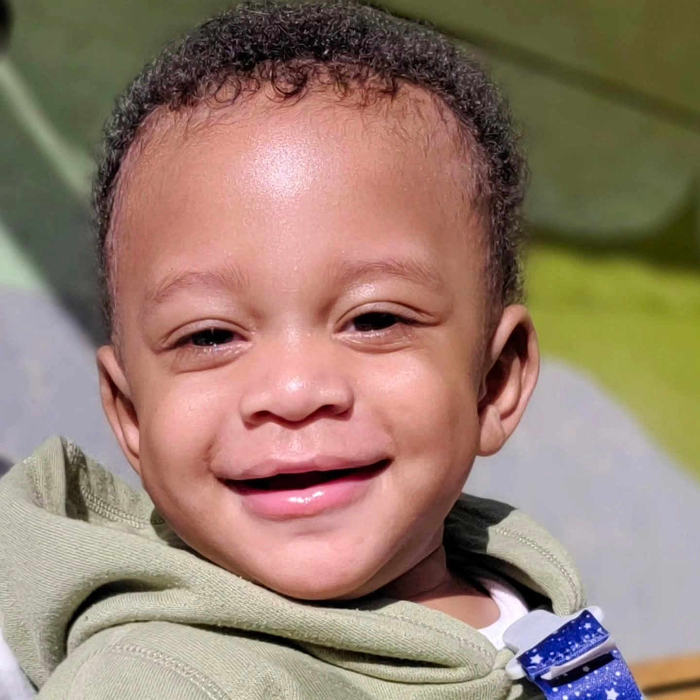 Issac is registered to the contest to win money with this photo: child, toddler, smiling, face, curly_hair, hoodie, closeup, happy, outdoor, sunlight, skin, young_child, portrait, person, baby, casual_clothing, cute, expression, daylight, infant