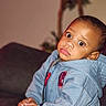child, toddler, hoodie, blue_clothing, indoor, couch, furniture, hands, face, curious_expression, person, young_child, casual_wear, home_interior, seated, portrait, brown_skin, short_hair, cute, baby