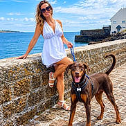 Mila participe au concours pour gagner de l'argent avec cette photo : woman, dog, doberman, outdoor, seaside, ocean, coast, promenade, stone_wall, stone_pavement, white_dress, sunglasses, leash, collar, standing, sitting, smiling, blue_sky, clouds, portrait