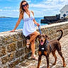 blue_sky, clouds, coast, collar, doberman, dog, leash, ocean, outdoor, portrait, promenade, seaside, sitting, smiling, standing, stone_pavement, stone_wall, sunglasses, white_dress, woman