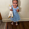 child, girl, toddler, portrait, smiling, happy, indoors, dress, blue_dress, hair_bows, braids, unicorn_plush, stuffed_animal, toy, sandals, standing, wood_floor, wall, birthday, fashion