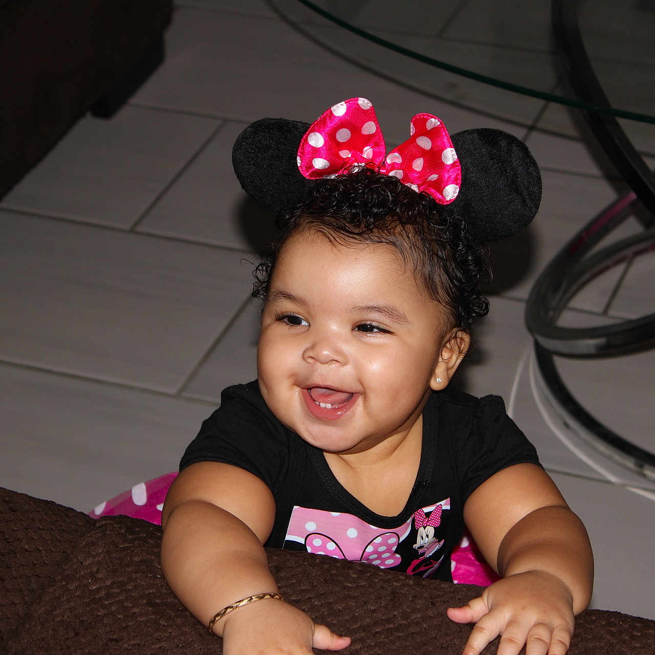 Amiry is registered to the contest to win money with this photo: toddler, child, smiling, minnie_mouse, headband, pink_bow, black_shirt, bracelet, curly_hair, indoor, glass_table, tiled_floor, happy, baby, person, cute, playful, joyful, furniture, home