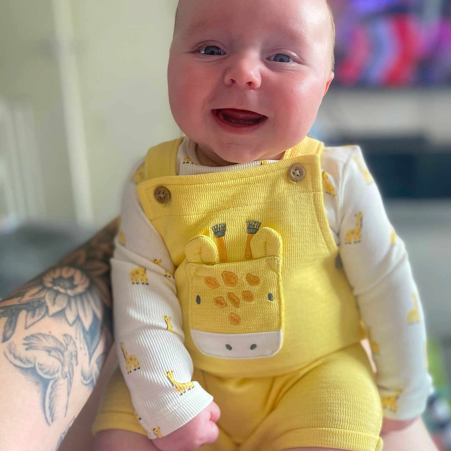 Oliver joined the competition — help win amazing prizes! arm, baby, blurred_background, child, closeup, clothing, cute, face, giraffe_design, happy, head, indoor, infant, living_room, person, sitting, skin, smiling, tattoo, yellow_outfit
