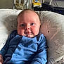 baby, infant, blue_onesie, cushion, patterned_fabric, face, chubby_cheeks, hand, high_chair, indoor, home, person, portrait, cute, child, seated, expression, furniture, skin, eyes