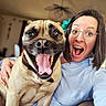 woman, dog, glasses, smile, tongue_out, indoor, happy, face, pet, collar, flower, hair, casual_clothing, furniture, hand, brown_dog, short_hair, portrait, excited, selfie
