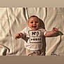 baby, smiling, infant, blanket, white_shirt, text_on_clothing, lying_down, happy, cute, indoor, child, person, clothing, patterned_pants, arms_outstretched, face, head, soft_texture, comfort, portrait