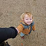 child, toddler, smile, holding_hand, adult_hand, jacket, hood, sand, beach, pebble, boots, shoes, sneakers, coat, outdoor, portrait, looking_up, happy, person, top_view