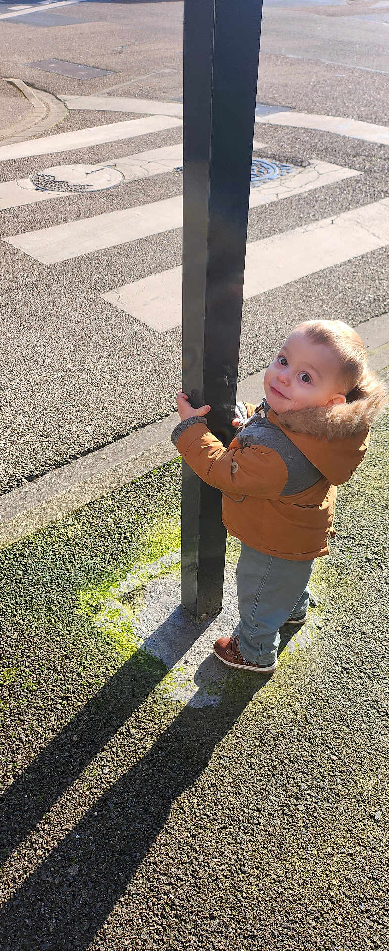 Aménadiel a rejoint le concours — aidez-le/la à gagner de superbes lots ! child, toddler, coat, hood, jacket, pole, street, crosswalk, pavement, shadow, shoe, sidewalk, standing, looking_up, outdoors, sunlight, moss, manhole, playful, portrait
